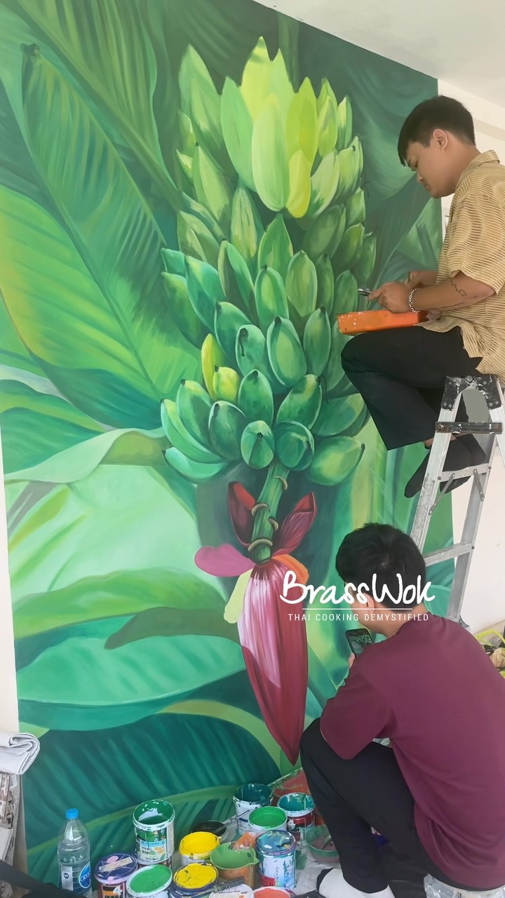 Three years ago when we re-opened BrassWok Thai Cooking Class after covid. A blank wall turned into a beautiful symbol of Thai abundance. ✨🎨 Watch as these two talented artists bring our mural to life here at BrassWok Thai Cooking Class Phuket!
Beyond being incredibly beautiful, this painting carries a deep meaning. In Thai culture, the banana plant and its blossom represent the sheer fertility and prosperity of our land. Thailand’s soil is so rich that these plants thrive naturally, blooming and offering food that never stops giving. 🍌🌿
It is the perfect symbol for our kitchen, where the love for Thai food is always blooming and always shared. Come see it in person during your next class!
#ThaiCookingClass #PrivateChefExperiecne #BrassWok #SupportLocalArtists #PhuketArt